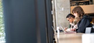 Students working on their laptops at a desk facing a window